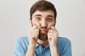 Close-up portrait of gloomy upset bearded male, squeezing cheeks with hands and looking at camera, being in bad mood