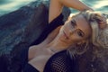 Close up portrait of gorgeous glam tanned blond woman wearing black swimsuit and summer tunic relaxing on the stones at the sea