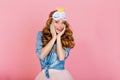 Close-up portrait of graceful curly girl in sleep mask and denim retro shirt, posing with hands touching face. Beautiful