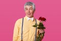 Close up portrait of grey haired elderly man in fashionable yellow shirt, suspenders, white bow tie, holds red roses, being in