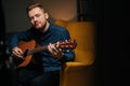 Close-up portrait of handsome guitarist singer male playing acoustic guitar sitting on armchair in dark living room. Royalty Free Stock Photo