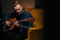 Close-up portrait of handsome guitarist singer male playing acoustic guitar sitting on armchair in dark living room. Royalty Free Stock Photo