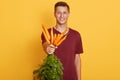 Close up portrait of handsome man holding bunch of fresh carrots isolated over yellow background, attractive guy wearing maroon Royalty Free Stock Photo