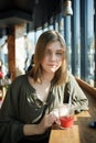 Close up portrait of happy beautiful teen student girl with a glass mug straw fruit tea at street cafe sitting near big window bit Royalty Free Stock Photo