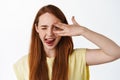 Close up portrait of happy cute girl with long red hiar, winking and showing tongue, open eye peek through fingers and