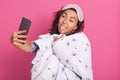 Close up portrait of happy female wrapped white blanket with dandelion, showing her tongue, making selfie, posing over