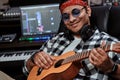 Close up portrait of happy young man, male artist in sunglasses smiling while playing ukulele, sitting in recording Royalty Free Stock Photo