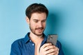 Close up portrait of happy young man reading mobile sreen, networking in smartphone, standing on blue background