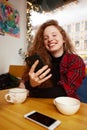 Close-up portrait of happy young woman making selfie on mobile phone sitting at table with laptop by window in cafe Royalty Free Stock Photo