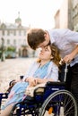 Close-up portrait of happy young woman in wheelchair and her husband kissing her forehead, walking outdoors in old city Royalty Free Stock Photo