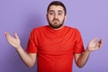 Close up portrait of helpless young handsome man raising hands, being disappoined, wearing red casual t shirt, standing isolated