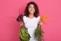 Close up portrait of hungry pleasant sweet brunette standing isolated over pink background, holding raw vegetables, having beet