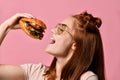 Close up portrait of a hungry young woman eating burger isolated over pink background