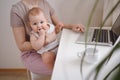 Close up portrait little baby boy funny facial expression sitting on mother`s lap studying laptop. Young mom work from home with