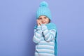 Close up portrait of little curly girl wearing warm winter hat, scarf and striped sweater, keeping hands near cheek, looking Royalty Free Stock Photo