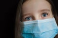 Close-up portrait of little defenseless girl on black backgound in blue disposable mask for protection of Coronavirus