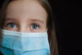 Close-up portrait of little defenseless girl on black backgound in blue disposable mask for protection of Coronavirus