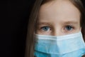 Close-up portrait of little defenseless girl on black backgound in blue disposable mask for protection of Coronavirus