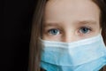 Close-up portrait of little defenseless girl on black backgound in blue disposable mask for protection of Coronavirus