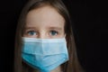 Close-up portrait of little defenseless girl on black backgound in blue disposable mask for protection of Coronavirus