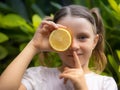Close up portrait of little girl with lemon. Covering eye with half of fresh lemon. Girl puts finger on lips, making hush sign or