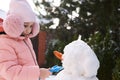 Close-up portrait lovely little child girl in pink down jacket and fluffy earmuffs, building snowman in snowy backyard Royalty Free Stock Photo