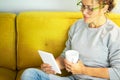 Close up portrait of mature pretty woman reading a book on tablet with glasses sitting on the yellow couch at home in break Royalty Free Stock Photo