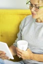 Close up portrait of mature pretty woman reading a book on tablet with glasses sitting on the yellow couch at home in break Royalty Free Stock Photo