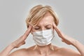 Close up portrait of middle aged woman wearing white medical mask, touching her temples, having headache, feeling stress