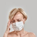 Close up portrait of middle aged woman wearing white medical mask, touching her temples, having headache, feeling stress