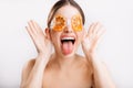 Close-up portrait of perky girl in eye mask with oranges showing tongue and making face on white background.