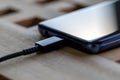 A close up portrait of a phone lying on a wooden table getting its battery charged using a USB cable connected to its port. The