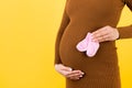 Close up portrait of pregnant woman in brown dress holding pink socks for a baby girl at yellow background. Childbirth expecting. Royalty Free Stock Photo