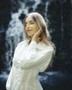 Close up portrait of pretty Caucasian woman near waterfall. Beautiful female with long blond hair wearing white dress. Closed eyes Royalty Free Stock Photo