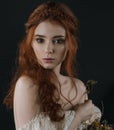 A close-up portrait of a red-haired young woman in a vintage gold dress with bare shoulders with a dry bouquet in hands on a black