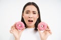 Close up portrait of sad asian woman, upset being on diet, showing two glazed pink doughnuts, tempted to eat junk food