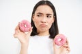 Close up portrait of sad asian woman, upset being on diet, showing two glazed pink doughnuts, tempted to eat junk food