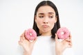 Close up portrait of sad asian woman, upset being on diet, showing two glazed pink doughnuts, tempted to eat junk food