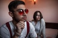 Close-up portrait of a sad brooding brunette guy in sunglasses. in the background on the bed a young brunette girl. selective