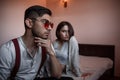 Close-up portrait of a sad brooding brunette guy in sunglasses. in the background on the bed a young brunette girl. selective