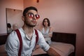 Close-up portrait of a sad brooding brunette guy in sunglasses. in the background on the bed a young brunette girl. selective