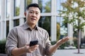 Close-up portrait of a sad young Asian man standing near an office building with a phone in his hands and looking Royalty Free Stock Photo