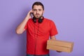 Close up portrait of serious bearded delivery man wearing red casual t shirt, talking via mobile phone with client, holding carton Royalty Free Stock Photo