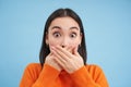 Close up portrait of shocked korean woman shuts her mouth with startled, impressed face expression, stands over blue