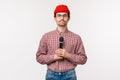 Close-up portrait of shy and geeky cute young bearded guy in glasses and red beanie, looking at tv screen and holding