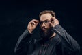 Close-up Portrait of a smart bearded guy in a black jacket corrects his glasses while standing in a dark studio