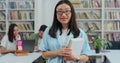 Close up portrait of smart beautiful young asian girl in glasses holding book in library bookshelf background looking at Royalty Free Stock Photo