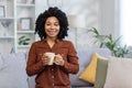 Close-up portrait of a smiling African American young woman relaxing on the couch at home, holding a cup of drink and Royalty Free Stock Photo