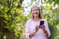 Close-up portrait of a smiling gray-haired sporty woman standing in the park wearing headphones, holding a phone and Royalty Free Stock Photo