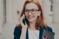 Smiling positive red-haired business woman wearing spectacles calling partner, standing outdoors